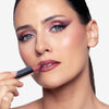 Close-up of a person applying matte pink lipstick, featuring dramatic purple eye shadow and winged eyeliner.