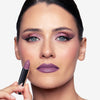 Woman showcasing lavender lipstick and shimmery eye makeup, holding a matching lipstick tube.