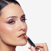 Woman applying nude lipstick, showcasing shimmering pink eyeshadow and precise winged eyeliner.