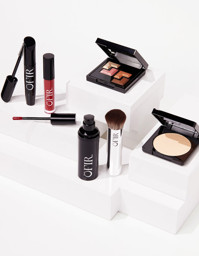 A selection of cosmetic products including a mascara, lip gloss, eyeshadow palette, face powder, and makeup brush arranged on white blocks.