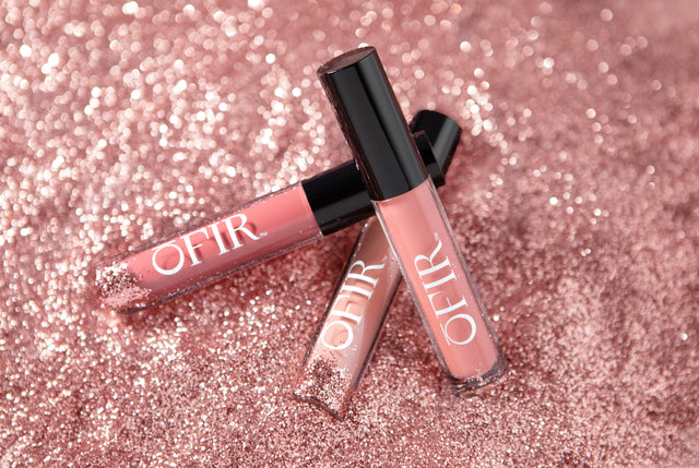 Three tubes of lip gloss with black caps labeled "OFIR" are arranged on a sparkling pink glitter surface.