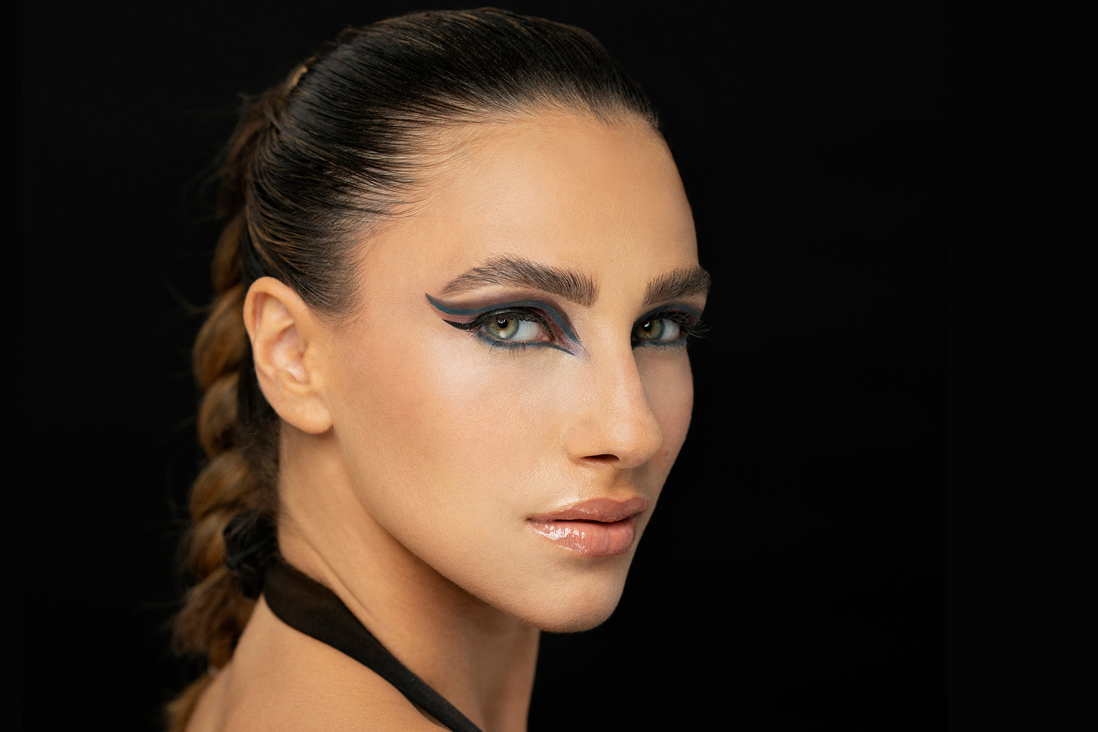 A person with dramatic winged eyeliner and glossy lips poses against a dark background, highlighting their makeup artistry.