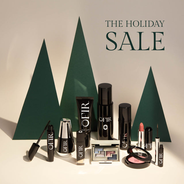 A collection of OFIR makeup products, including mascara, lipstick, eyeshadow, and foundation, is displayed against a backdrop of green paper trees with "The Holiday Sale" text.