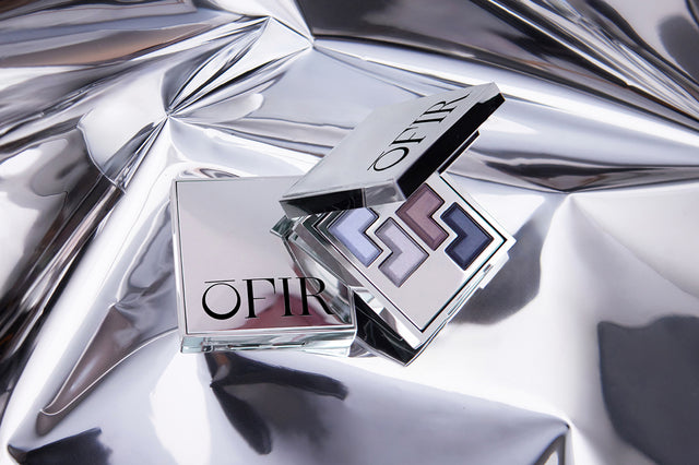 Open silver compact showcasing a set of four purple and blue eyeshadows, set against a reflective metallic background.