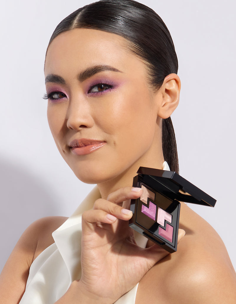 A woman with purple eyeshadow and glossy lips holds up a compact with pink and beige eyeshadow shades, wearing a sleek, white garment.