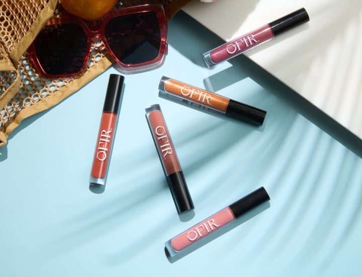 Five OFIR liquid lip products in warm terracotta, nude, orange, and berry shades on a light blue surface with sunglasses and a mesh bag