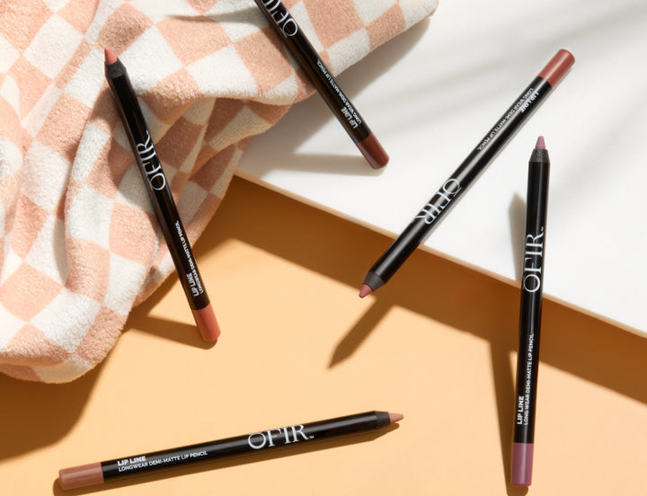 Five OFIR Lip Line long-wear demi-matte lip pencils in nude and mauve shades scattered on a peach and white checkered towel surface