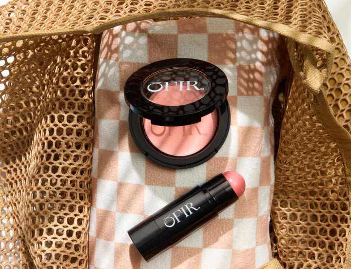 OFIR blush compact and matching lip color stick in coral pink displayed inside a woven straw bag on a checkered beach towel